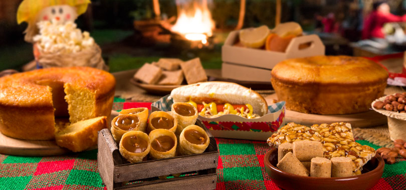 Comidas de festa junina para transformar seu cardápio no mês de junho 2 Mesa com comidas de festa junina, como bolos, paçoca, cachorro-quente e canudos de doce de leite.