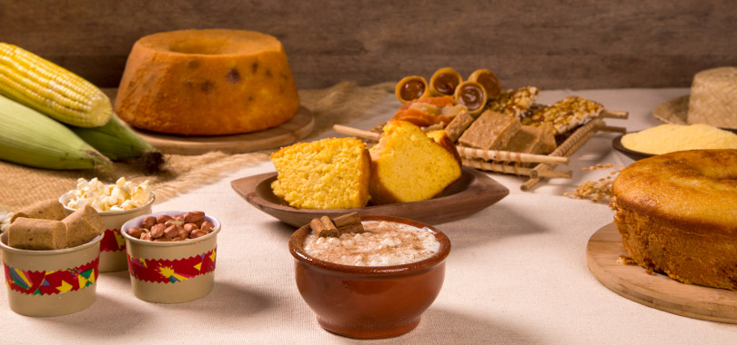 Mesa com comida de festa junina, como bolo de fubá com goiabada, espiga de milho, canjica e canudos de doce de leite.