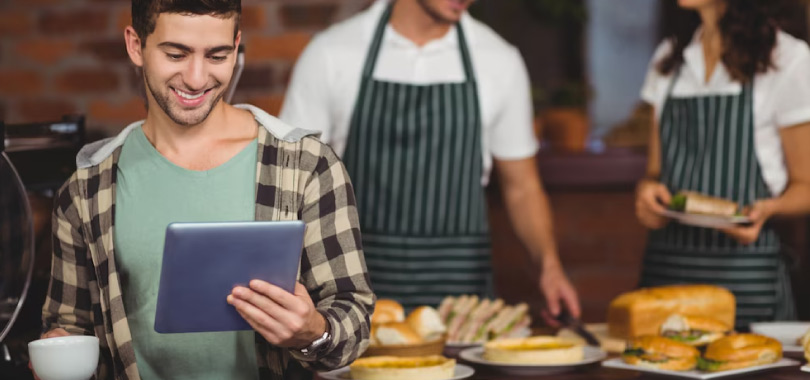 Conheça as melhores estratégias de fidelização de clientes para o setor de Food Service 2 Funcionário sorrindo com tablet na mão, ao fundo outros dois funcionários.
