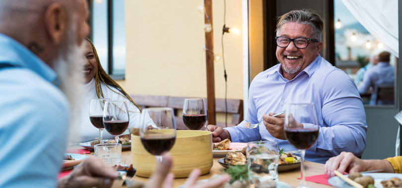 Crie um cardápio de Dia dos Pais especial e atraia mais clientes 2 Pais sorrindo em almoço em restaurante. Mesas com comidas variadas e taças de vinho tinto.