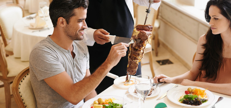 Casal almoçando em churrascaria. Homem está pegando pedaço de carne do espeto.