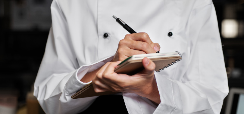 Pessoa, usando avental branco, anotando em caderno.