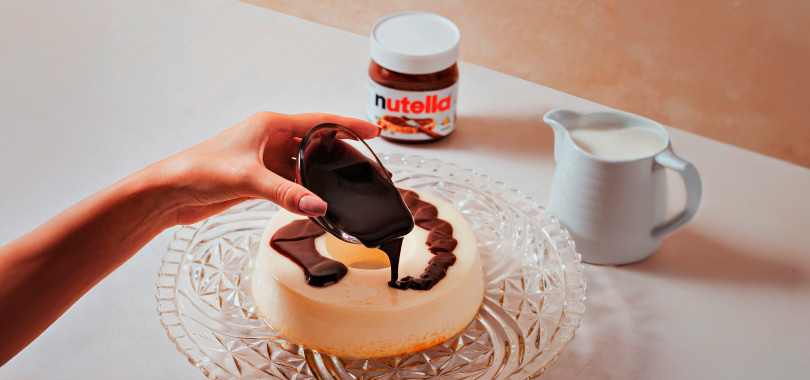 Ideias para comemorar o Dia Mundial da Nutella com seus clientes 2 Mão despejando calda de Nutella em cima de pudim.