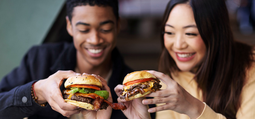 Aproveite o Dia Mundial do Hambúrguer para alavancar as vendas do seu empreendimento 2 Homem e mulher segurando dois hambúrgueres.