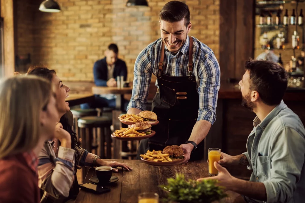 Garçom sorridente servindo hambúrgueres e batatas fritas a grupo de amigos em pub aconchegante, ideia para Black Friday.